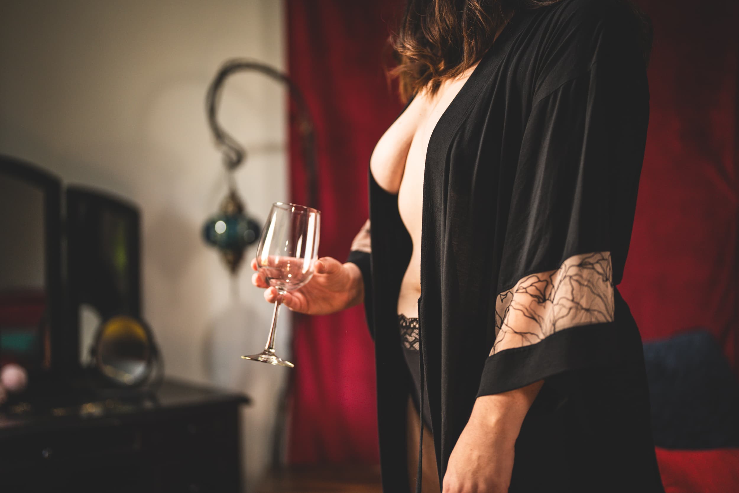 A woman in a black robe and lingerie, holding a wine glass in one hand, with a vintage interior background.