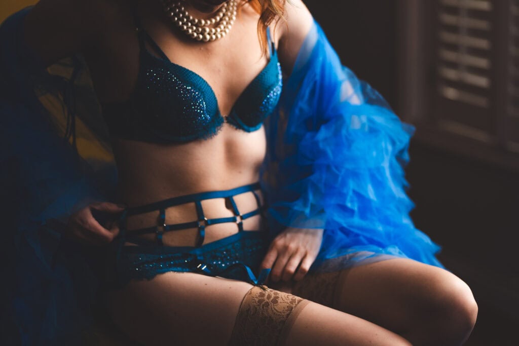 Close-up of a woman in a sparkling blue sequined lingerie set with a dramatic blue tulle wrap and intricate straps around her midriff. She accessorizes with a pearl necklace, seated in a relaxed yet confident pose, against a softly lit, moody background.